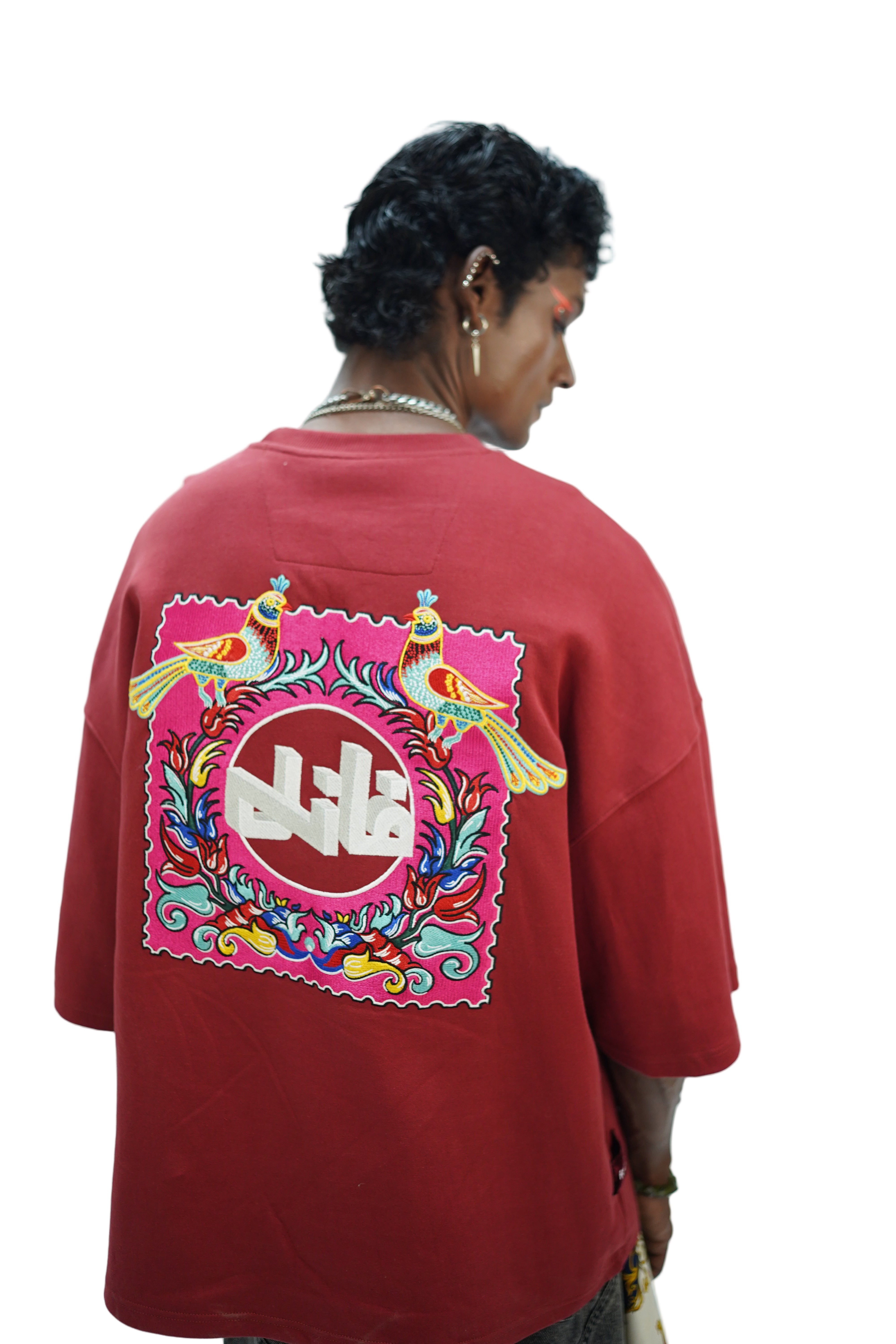 Model wearing oversized maroon FAANI embroidered t-shirt featuring two colorful birds and a bold Urdu logo framed by floral truck-art inspired motifs on the back