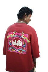 Model wearing oversized maroon FAANI embroidered t-shirt featuring two colorful birds and a bold Urdu logo framed by floral truck-art inspired motifs on the back